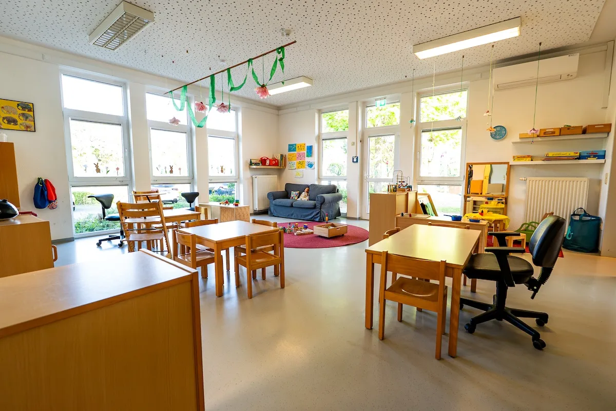 Heller Gruppenraum der Nestgruppe mit großen Fenstern, blauem Sofa, rotem Spielteppich, Kindertischen und bunter Kinderkunst in der Kita Schweich