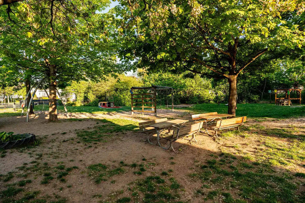 Außengelände der Kita Schweich mit Spielplatz, großen Bäumen und Sitzbänken